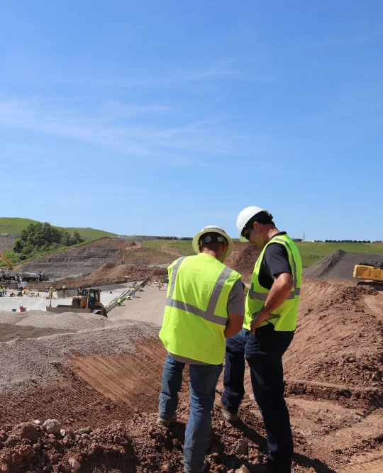 Two workers in safety gear on a project site consulting project plans