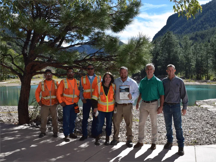 Four workers in safety gear and three individuals in business casual wear standing together in front of the lake and trees, holding an award