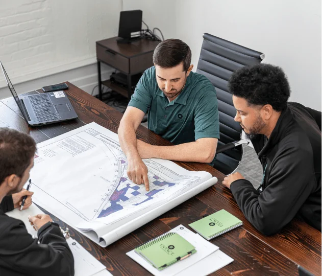 Three individuals in an office looking at project plans on a large wooden desk