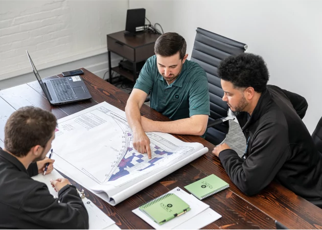 Three individuals in an office looking at project plans on a large wooden desk