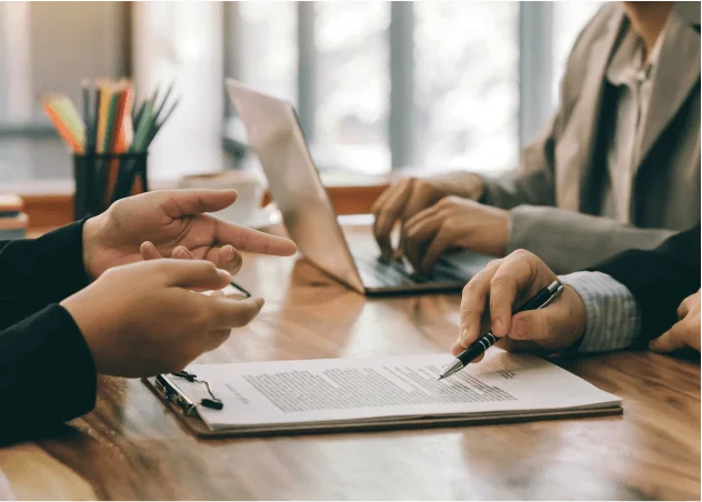 Close-up of two individuals at a desk reviewing documents. Another individual is typing on a laptop in the background