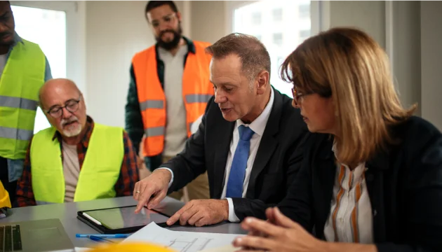 A team of employees in safety gear and business professionals discussing plans in an office looking at a tablet