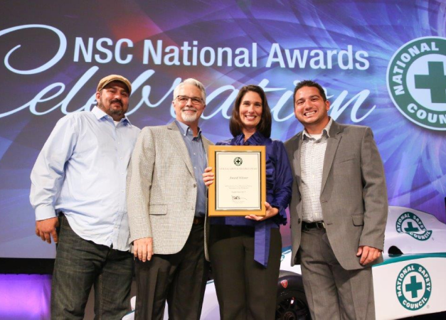 Four individuals in business professional clothing standing together on a stage holding a National Safety Council award