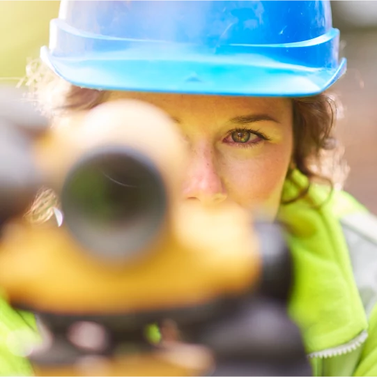 Worker looking through surveyor equipment with blue hardhat on